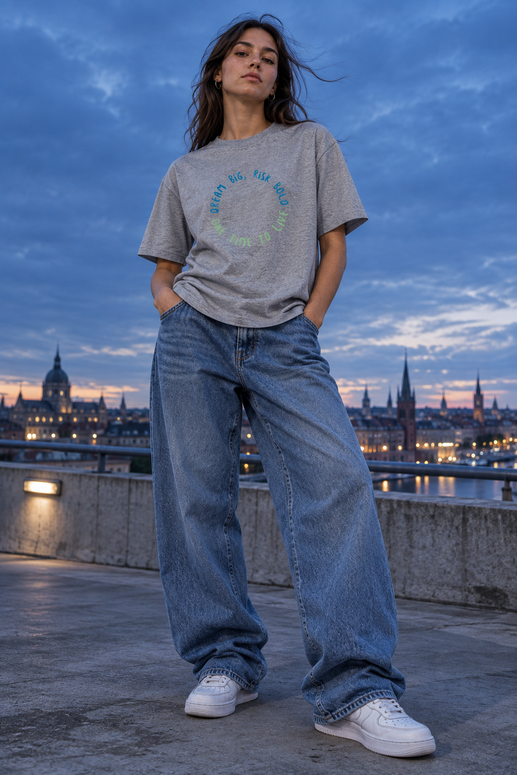 Woman wearing the 1TIME2 Dream Big organic tee in Heather Grey with wide-leg jeans, city skyline at dusk